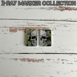May include: Two small, square, white x-ray markers with a camouflage pattern and a skull graphic. The text "RAY" is visible on each marker.  The markers are on a white wooden surface.  Camo Split Skull X-Ray Marker Collection.