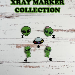 May include: A set of five green alien-themed x-ray markers. The markers are numbered 1 through 5. The markers feature different alien designs, including a UFO, an alien with a peace sign, and an alien with a bow. The text "I NEED SPACE XRAY MARKER COLLECTION" is at the top of the image. The text "ADD PIXIE GLITTER, NO EXTRA CHARGE" is at the bottom of the image.