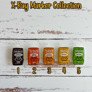 May include: Five colorful enamel pins shaped like hot sauce packets with cat illustrations and text. The pins are labeled 1 through 5 and the text on the pins reads "Diablo", "Fire", "Hot", "Mild", and "Verde". The text below the pins reads "add pixie glitter".