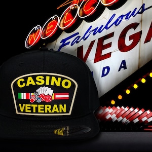 May include: Black baseball cap with a patch that reads "CASINO VETERAN." The patch features playing cards, dice, and flags. The background includes the "Welcome to Fabulous Las Vegas Nevada" sign and casino chips.