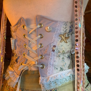 May include: A lavender corset with gold eyelets and white lacing. The corset features floral fabric panels, lace trim, and decorative seashell and starfish embellishments. A gold chain and ribbon accents add detail to this garment.