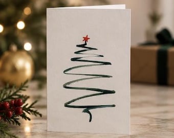 May include: A white greeting card featuring a hand-painted dark green watercolor brushstroke forming a stylized Christmas tree shape. A small, bright red star is painted at the very top of the tree, completing the simple, minimalist design.