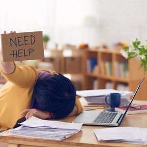 Puede incluir: Una persona sentada en un escritorio con un ordenador portátil, papeles y una taza de café. Está sosteniendo un cartel de cartón que dice "NEED HELP".
