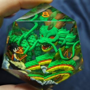 Puede incluir: Un dado grande de resina transparente de múltiples caras con un dragón verde con ojos rojos y un vientre amarillo y rojo. También se ve una pequeña figura. El dado tiene números naranjas.