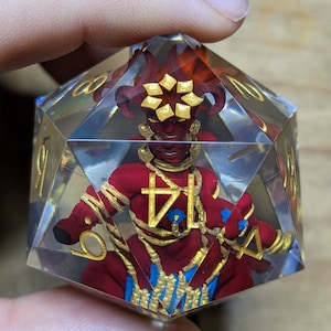 Puede incluir: Un dado transparente de múltiples caras con una figura roja y dorada en su interior. La figura tiene cuernos, una cara roja y lleva joyas doradas. El dado tiene números dorados en cada lado.