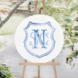 May include: A round white sign with a blue shield design and the letters "SM" and "N" in a decorative font. The sign is displayed on a wooden easel outdoors, with greenery in the background.