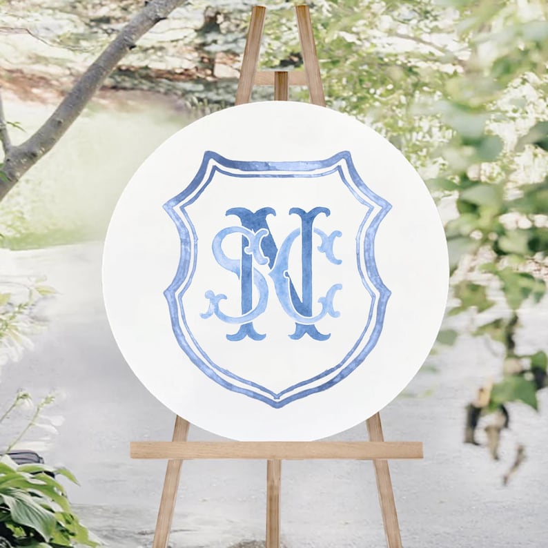 May include: A round white sign with a blue shield design and the letters "SM" and "N" in a decorative font. The sign is displayed on a wooden easel outdoors, with greenery in the background.