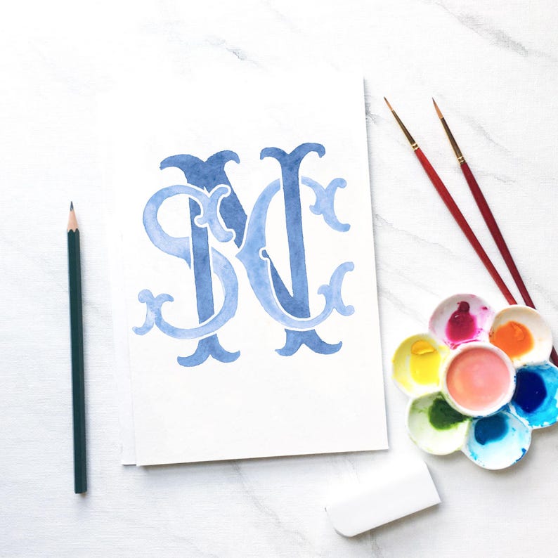 May include: Watercolor painting of the intertwined letters "SN" in shades of blue on white paper. A dark green pencil, two paintbrushes, a white eraser, and a palette of colorful paints are arranged around the artwork.
