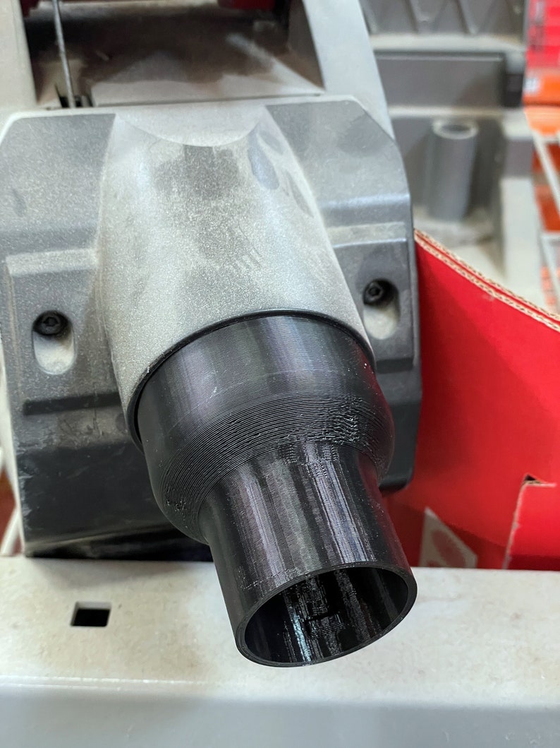 May include: A black plastic adapter attached to a grey power tool. The adapter has a cylindrical shape with a flared opening. The tool appears to be a saw, with a red cardboard box visible in the background.
