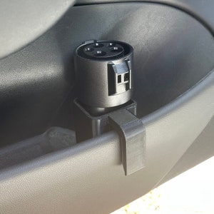 K&ouml;nnte beinhalten: Ein schwarzer Ladeport-Halter f&uuml;r ein Elektrofahrzeug, der an einer Autot&uuml;rverkleidung befestigt ist. Der Halter dient zur Sicherung des Ladeanschlusses und h&auml;lt ihn zug&auml;nglich und organisiert. Der Halter besteht aus schwarzem Kunststoff.