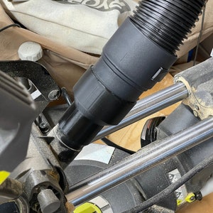 K&ouml;nnte beinhalten: Ein schwarzer Staubsaugerschlauch, der an einer Gehrungss&auml;ge befestigt ist. Der Schlauch ist gewellt und mit einem schwarzen Adapter verbunden. Die S&auml;ge hat silberne Metallstangen und eine gelb-schwarze Basis. S&auml;gemehl ist sichtbar.