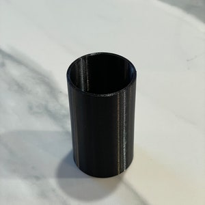 May include: A black cylindrical object with a hollow interior. The object is made of a solid material and has a smooth, matte finish. The cylinder is standing upright on a white surface.