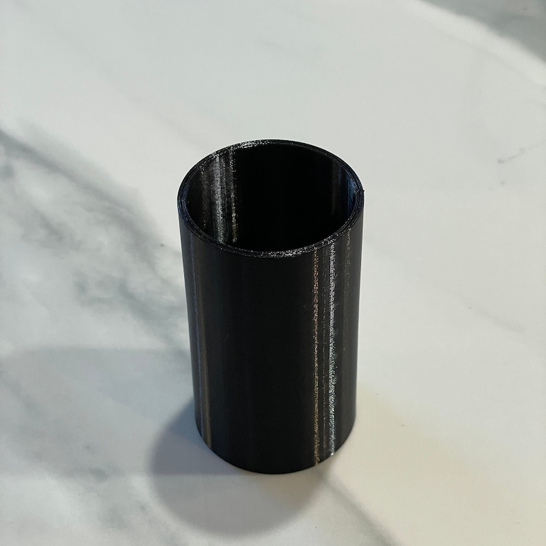 May include: A black cylindrical object with a hollow interior. The object is made of a solid material and has a smooth, matte finish. The cylinder is standing upright on a white surface.