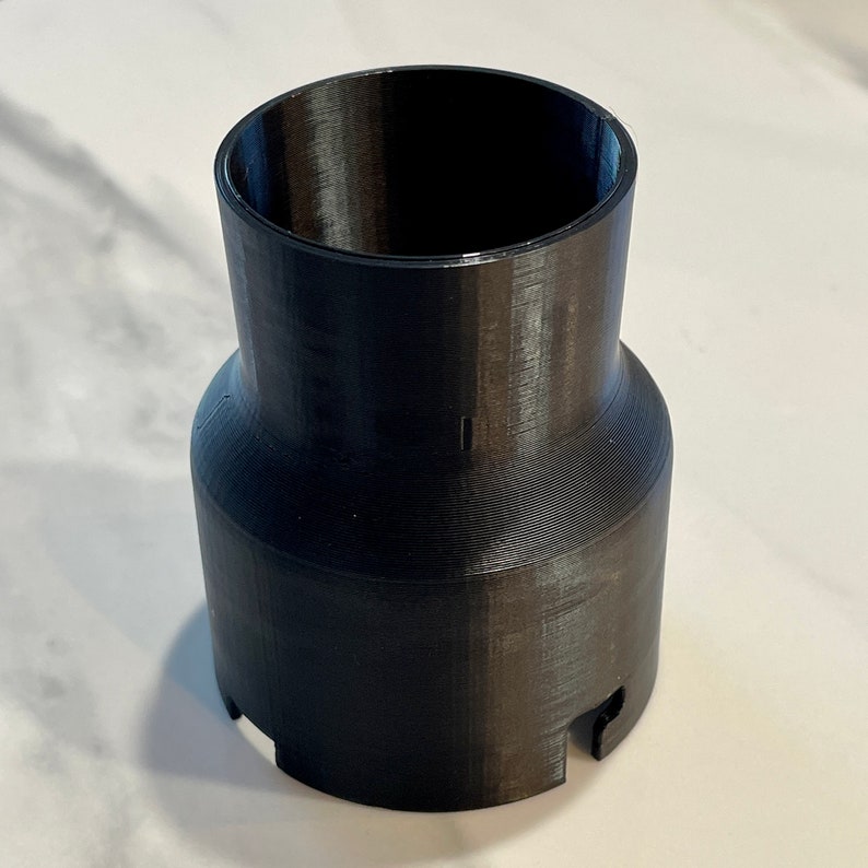 May include: A black, cylindrical 3D-printed object with a flared top and a wider base. The object has a ridged texture and two small cutouts near the base. The interior is hollow, and the overall shape is a combination of a cylinder and a cone.