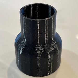 May include: A black cylindrical adapter with a flared base and a narrower top. The adapter appears to be 3D-printed, with visible layer lines. The object is designed to connect two different sized pipes or fittings.