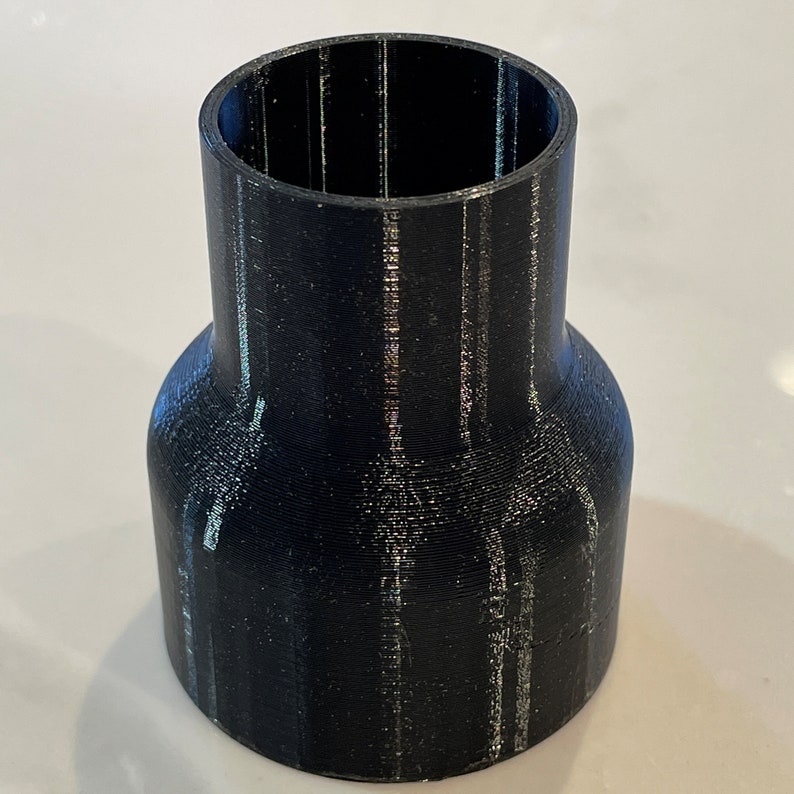 May include: A black cylindrical adapter with a flared base and a narrower top. The adapter appears to be 3D-printed, with visible layer lines. The object is designed to connect two different sized pipes or fittings.