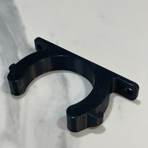 May include: A black 3D-printed bracket with a curved holder and a flat mounting surface. The bracket has two holes for screws and is designed to hold an object. The material has a textured surface.
