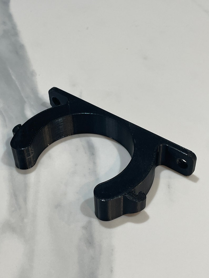 May include: A black 3D-printed bracket with a curved holder and a flat mounting surface. The bracket has two holes for screws and is designed to hold an object. The material has a textured surface.