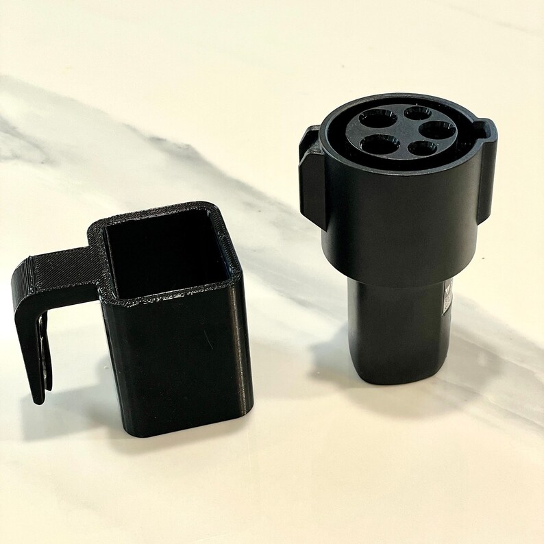K&ouml;nnte beinhalten: Zwei schwarze Komponenten zum Aufladen von Elektrofahrzeugen. Einer ist ein Ladeanschluss mit f&uuml;nf L&ouml;chern, der andere ein quadratischer Adapter mit Griff. Auf einer wei&szlig;en Oberfl&auml;che.