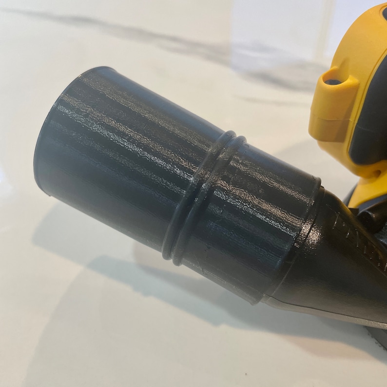 Puede incluir: Un accesorio cil&iacute;ndrico negro para una herramienta el&eacute;ctrica. El accesorio tiene una superficie texturizada y dos anillos elevados cerca del centro. Una parte de una herramienta el&eacute;ctrica amarilla y negra es visible en el fondo.