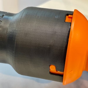 May include: Close-up of a black cylindrical object with a textured surface and an orange component. The orange piece has a small button and a triangular marking. The object appears to be made of plastic.