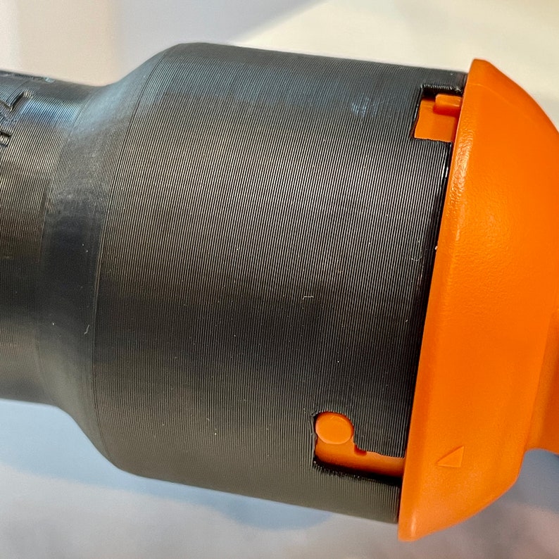 May include: Close-up of a black cylindrical object with a textured surface and an orange component. The orange piece has a small button and a triangular marking. The object appears to be made of plastic.