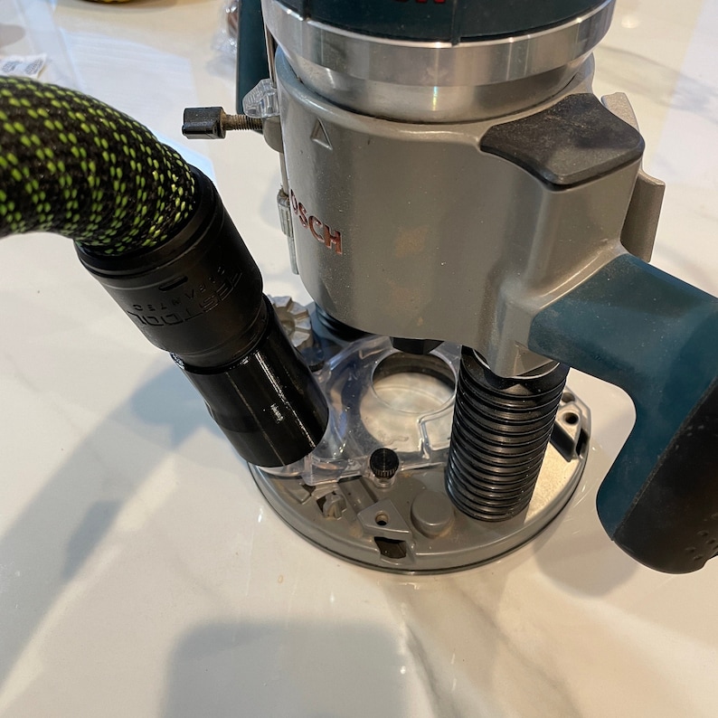 May include: A gray and teal Bosch router with a black and green hose attached. The router has a clear plastic base and a silver metal plate. The hose is connected to a black adapter.