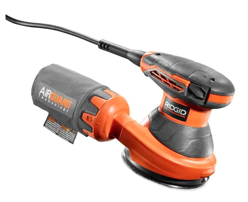 May include: An orange and black Ridgid random orbit sander with a black power cord. The sander has a round base and a dust collection bag with "AIR GUARD TECHNOLOGY" printed on it. The Ridgid logo is visible on the top of the sander.