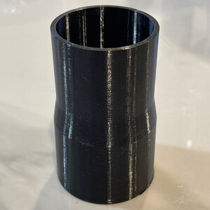 May include: A black 3D-printed cylindrical adapter with a flared top and bottom. The adapter has a smooth, matte finish and is designed to connect pipes or tubes of different sizes. The object is made of plastic.