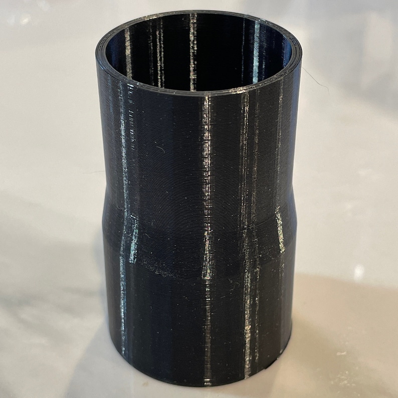 May include: A black 3D-printed cylindrical adapter with a flared top and bottom. The adapter has a smooth, matte finish and is designed to connect pipes or tubes of different sizes. The object is made of plastic.