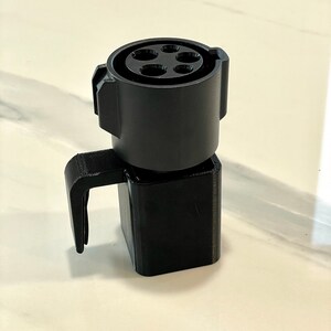 K&ouml;nnte beinhalten: Ein schwarzer EV-Ladeadapter mit Griff. Der Adapter hat eine kreisf&ouml;rmige Oberseite mit f&uuml;nf L&ouml;chern und eine quadratische Basis. Der Griff ist an der Basis befestigt und erstreckt sich nach au&szlig;en. Der Adapter besteht aus Kunststoff.