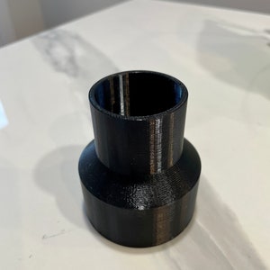 K&ouml;nnte beinhalten: Ein schwarzer, 3D-gedruckter Kunststoffadapter mit zylindrischem Oberteil und breiterer, konischer Basis. Der Adapter dient zum Verbinden von Komponenten unterschiedlicher Gr&ouml;&szlig;e. Die Oberfl&auml;che ist glatt und matt.