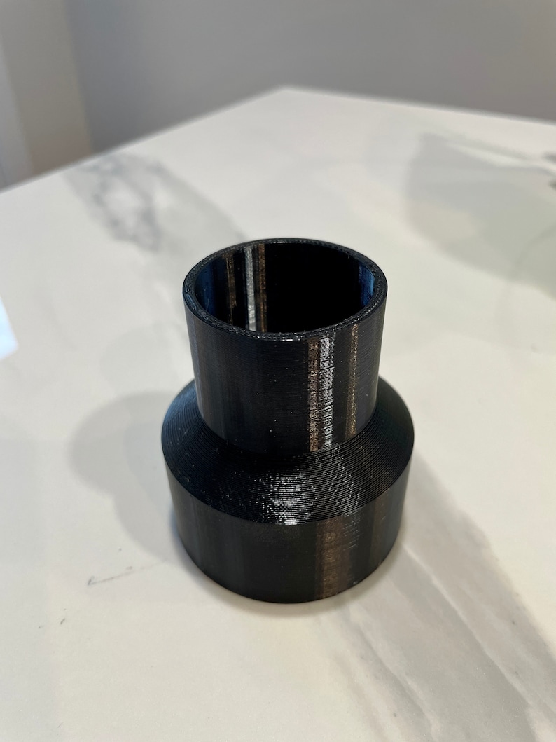 K&ouml;nnte beinhalten: Ein schwarzer, 3D-gedruckter Kunststoffadapter mit zylindrischem Oberteil und breiterer, konischer Basis. Der Adapter dient zum Verbinden von Komponenten unterschiedlicher Gr&ouml;&szlig;e. Die Oberfl&auml;che ist glatt und matt.