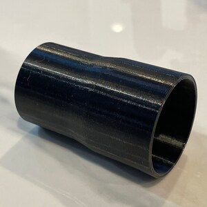 May include: A black cylindrical object with a slightly curved middle section. The object has a hollow interior and a textured surface, likely 3D-printed. The ends are open, revealing the interior. The overall shape is a short tube.