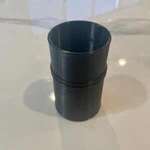 Puede incluir: Objeto cil&iacute;ndrico gris oscuro impreso en 3D con una superficie ligeramente texturizada. Tiene una base m&aacute;s ancha y una parte superior m&aacute;s estrecha, con una banda en relieve en el medio. La parte superior abierta sugiere un contenedor.