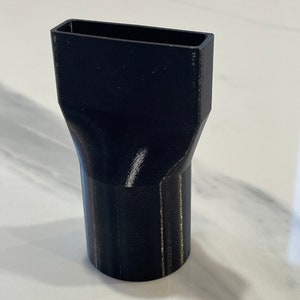 以下が含まれることがあります： 長方形の開口部と先細りのデザインが特徴の、黒色の3Dプリントノズルアタッチメント。ノズルは、おそらく気流の方向を変えたり、材料を分配したりするために、より大きなデバイスに取り付けるように設計されています。このオブジェクトは、固くて暗い素材で作られています。