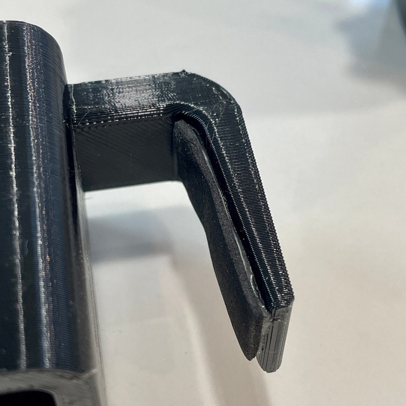K&ouml;nnte beinhalten: Nahaufnahme eines schwarzen 3D-gedruckten Objekts, m&ouml;glicherweise ein Griff oder eine Haltestruktur. Das Objekt hat eine zylindrische Basis und einen rechtwinkligen Ansatz mit einer flachen, strukturierten Oberfl&auml;che. Die Schichten des 3D-Druckprozesses sind sichtbar.