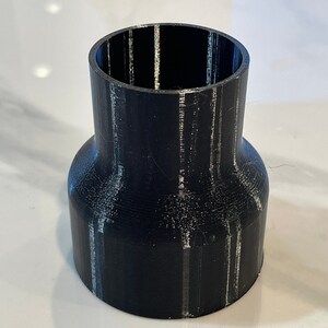 May include: A black 3D-printed pipe fitting with a flared base and a narrower top opening. The fitting has a cylindrical shape with visible layer lines from the printing process. The object is designed to connect pipes.