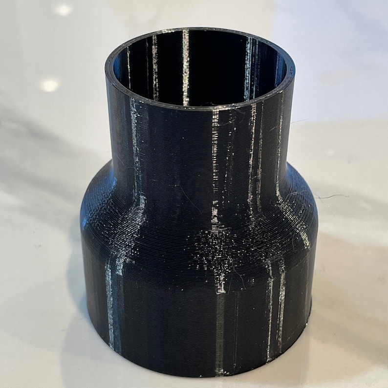 May include: A black 3D-printed pipe fitting with a flared base and a narrower top opening. The fitting has a cylindrical shape with visible layer lines from the printing process. The object is designed to connect pipes.