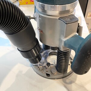 May include: A silver and teal Bosch router with a black hose attached. The router has a clear plastic base and a black handle. The Bosch logo is visible on the side of the tool.