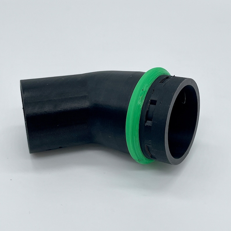 May include: A black, curved automotive part with a green circular seal. The part has a textured surface and a cylindrical opening on one end. The green seal is a bright, contrasting color against the black.