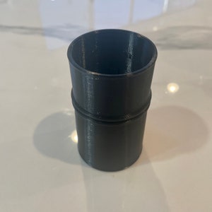 May include: A cylindrical, dark gray 3D-printed object with a slightly textured surface. The object has a wider base and a narrower top, with a raised band in the middle. The open top suggests it could be a container or a part of a larger assembly.