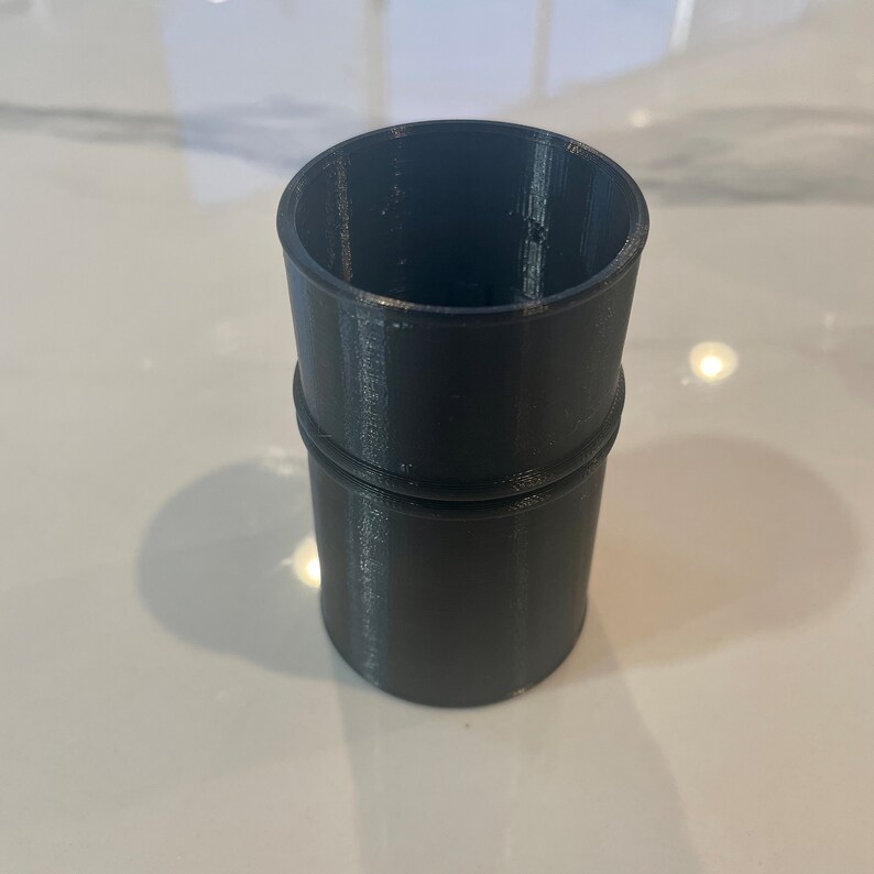 May include: A cylindrical, dark gray 3D-printed object with a slightly textured surface. The object has a wider base and a narrower top, with a raised band in the middle. The open top suggests it could be a container or a part of a larger assembly.