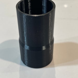 K&ouml;nnte beinhalten: Ein schwarzer, zylindrischer Adapter mit breiterem oberen und schmalerem unteren Teil. Die Oberfl&auml;che ist strukturiert, mit sichtbaren Schichtlinien, was auf 3D-Druck hindeutet. Er dient zum Verbinden von Komponenten unterschiedlicher Gr&ouml;&szlig;e.