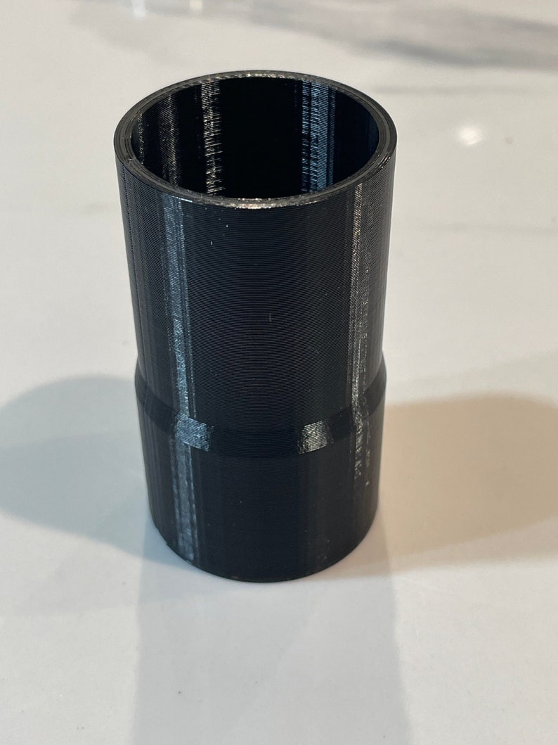 K&ouml;nnte beinhalten: Ein schwarzer, zylindrischer Adapter mit breiterem oberen und schmalerem unteren Teil. Die Oberfl&auml;che ist strukturiert, mit sichtbaren Schichtlinien, was auf 3D-Druck hindeutet. Er dient zum Verbinden von Komponenten unterschiedlicher Gr&ouml;&szlig;e.