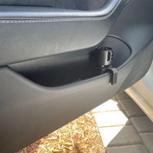 K&ouml;nnte beinhalten: Ein schwarzer Ladeadapter f&uuml;r ein Elektrofahrzeug, der in der T&uuml;rablage eines Autos befestigt ist. Der Adapter ist zylindrisch mit mehreren Stiftanschl&uuml;ssen. Die Autot&uuml;r ist schwarz mit wei&szlig;en N&auml;hten.