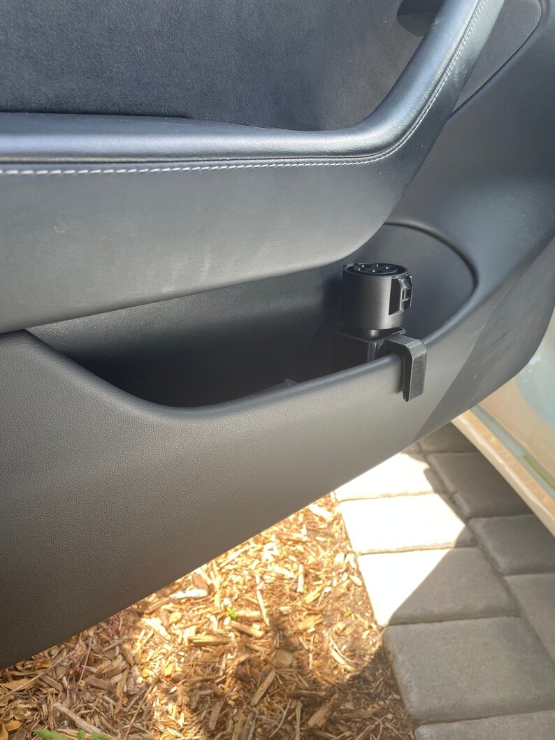 K&ouml;nnte beinhalten: Ein schwarzer Ladeadapter f&uuml;r ein Elektrofahrzeug, der in der T&uuml;rablage eines Autos befestigt ist. Der Adapter ist zylindrisch mit mehreren Stiftanschl&uuml;ssen. Die Autot&uuml;r ist schwarz mit wei&szlig;en N&auml;hten.