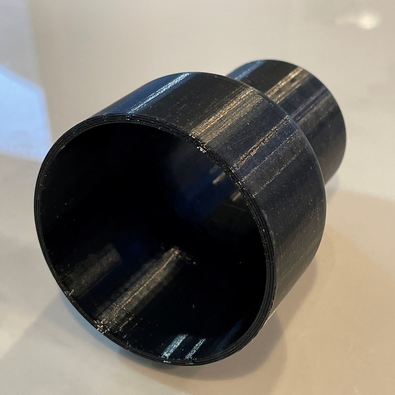 Puede incluir: Un objeto negro, cil&iacute;ndrico, impreso en 3D, con una abertura ensanchada y una extensi&oacute;n cil&iacute;ndrica m&aacute;s peque&ntilde;a. El objeto tiene una superficie texturizada y parece estar hecho de pl&aacute;stico. El dise&ntilde;o sugiere un conector o adaptador.