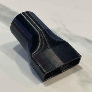 以下が含まれることがあります： 円筒形から長方形の開口部に移行する黒色の3Dプリントアダプター。 アダプターは角度がついており、テクスチャード加工されたプラスチック素材でできているようです。 オブジェクトは白い表面に配置されています。