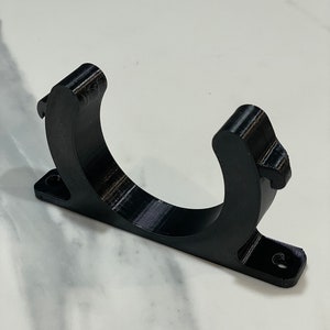 May include: A black, 3D-printed bracket with a curved cradle and a rectangular base. The base has two holes for mounting. The bracket is designed to hold a cylindrical object. The material has visible layer lines.